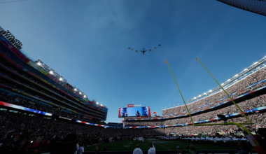A stadium filled with spectators watches a formation of military planes flying overhead during a daytime event.