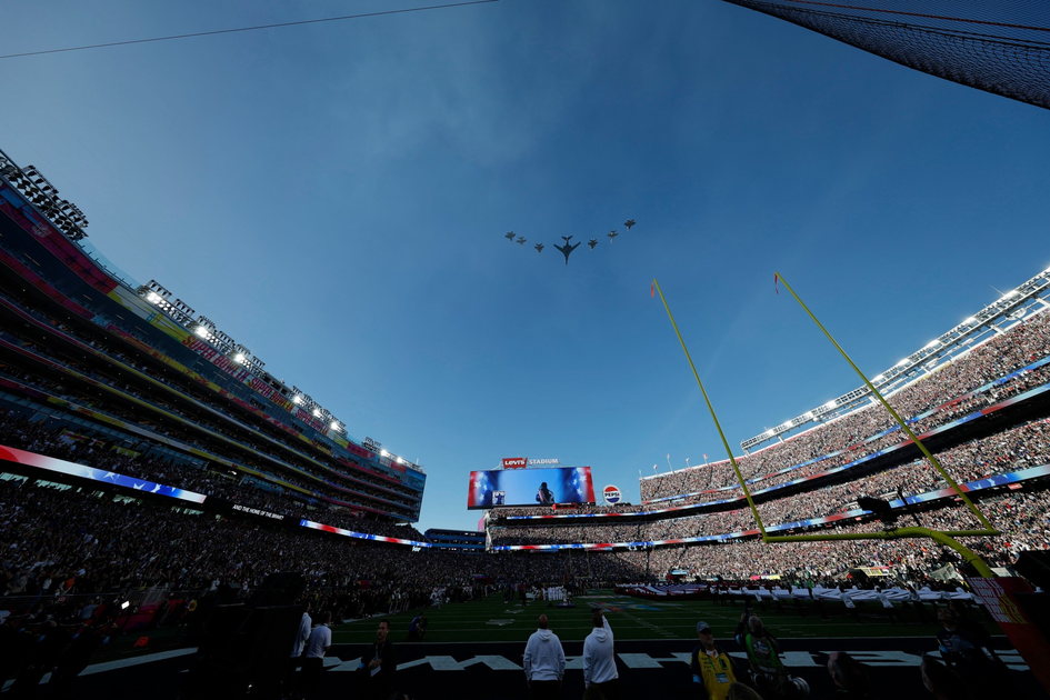 A stadium filled with spectators watches a formation of military planes flying overhead during a daytime event.
