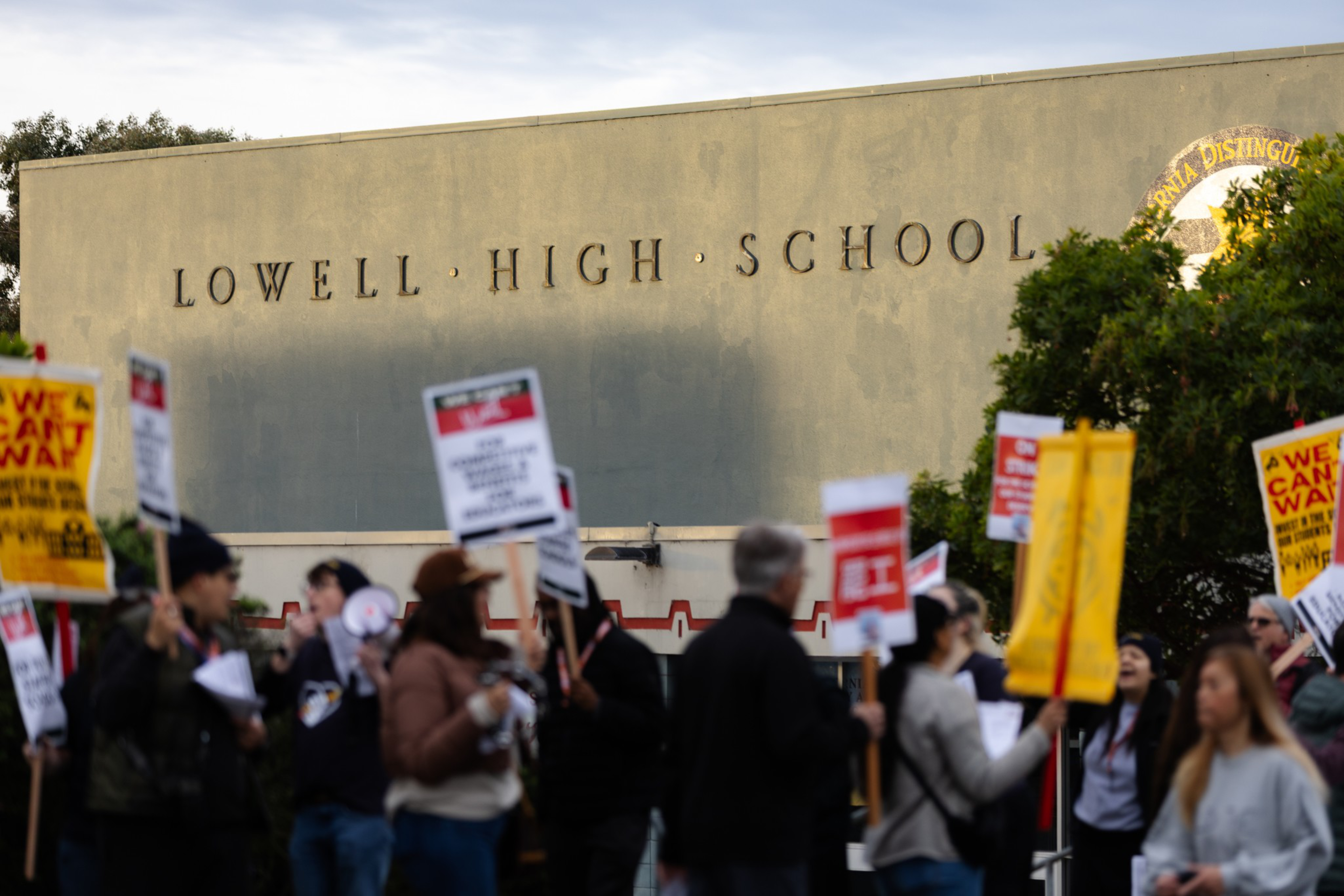 People holding protest signs gather outside a building labeled “Lowell High School” under a clear sky.