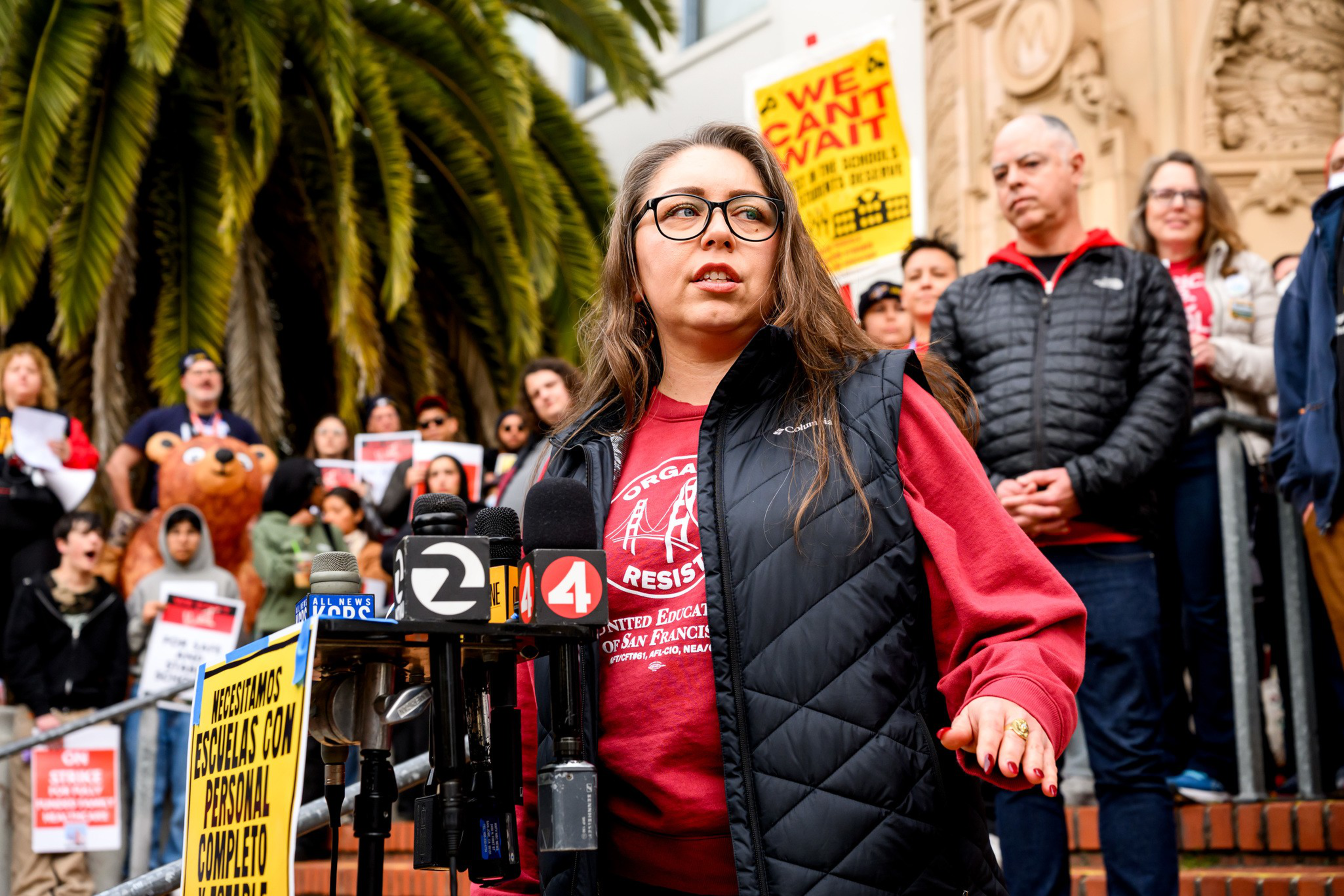 A woman wearing glasses and a red sweatshirt speaks at a protest with microphones nearby and people holding signs behind her.