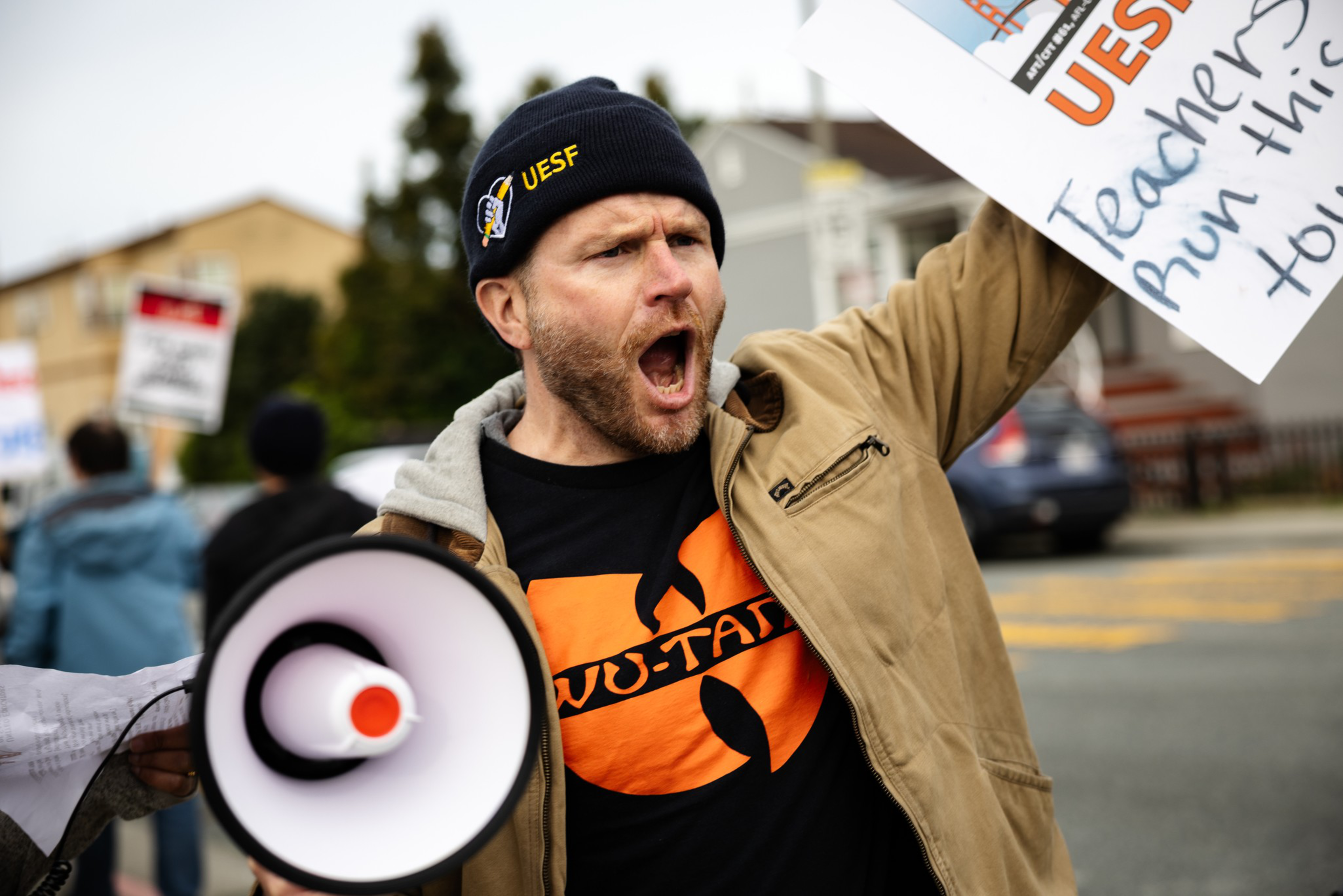 A man wearing a UESF beanie and Wu-Tang t-shirt holds a megaphone and a sign, shouting passionately during a street protest.