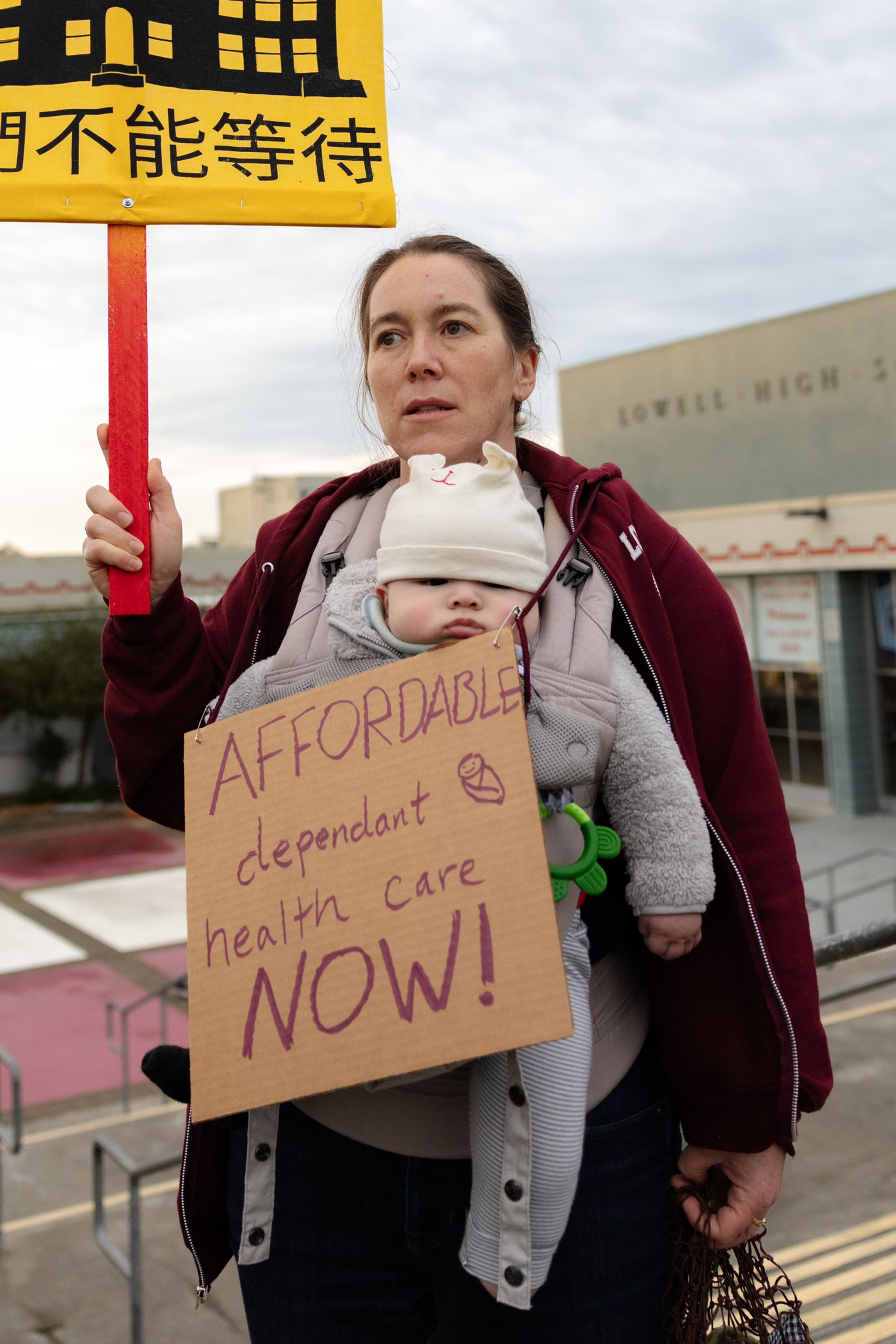 A woman carries a baby in a front carrier with a cardboard sign that reads “AFFORDABLE dependant health care NOW!” and holds a yellow protest sign with Chinese characters.