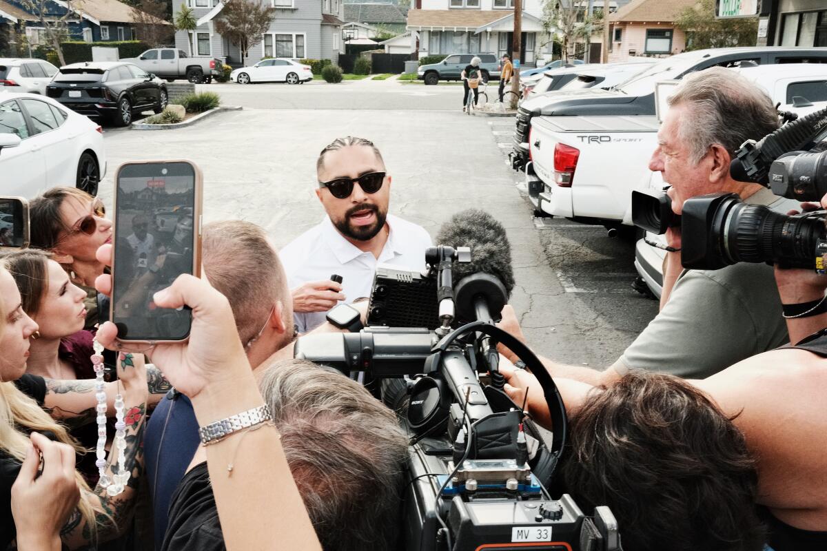A man in sunglasses takes questions from press in a parking lot