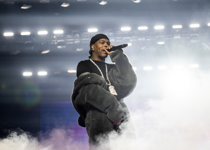 A performer wearing a dark beanie, oversized furry jacket, and a large chain necklace holds a microphone, surrounded by stage fog and bright lights.