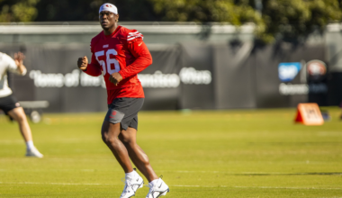 A football player in a red jersey numbered 56 and black shorts runs on a green field during practice with trees and a banner in the background.