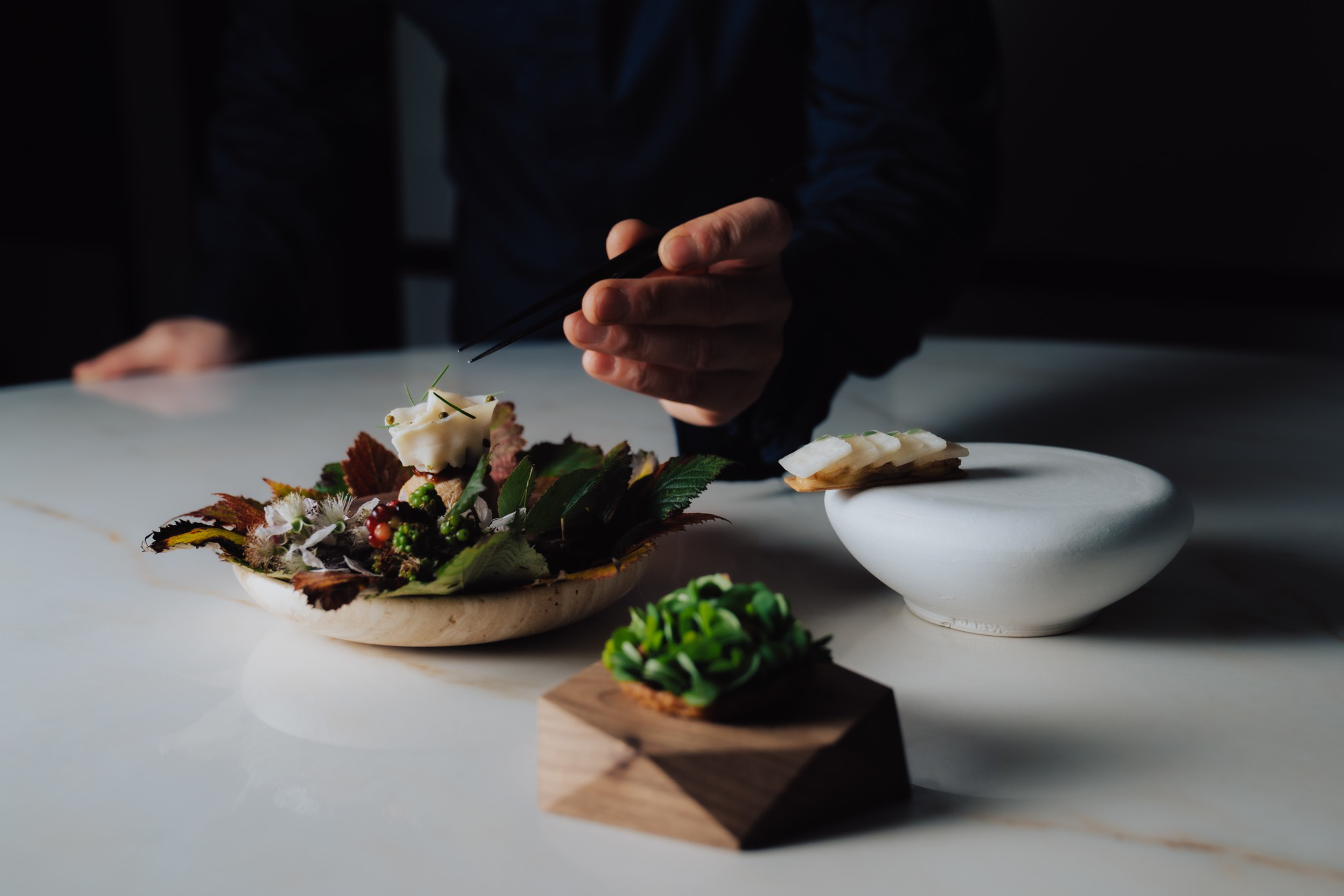 A person uses tweezers to delicately place an ingredient on a dish with leaves and flowers, beside two artfully presented small food items.