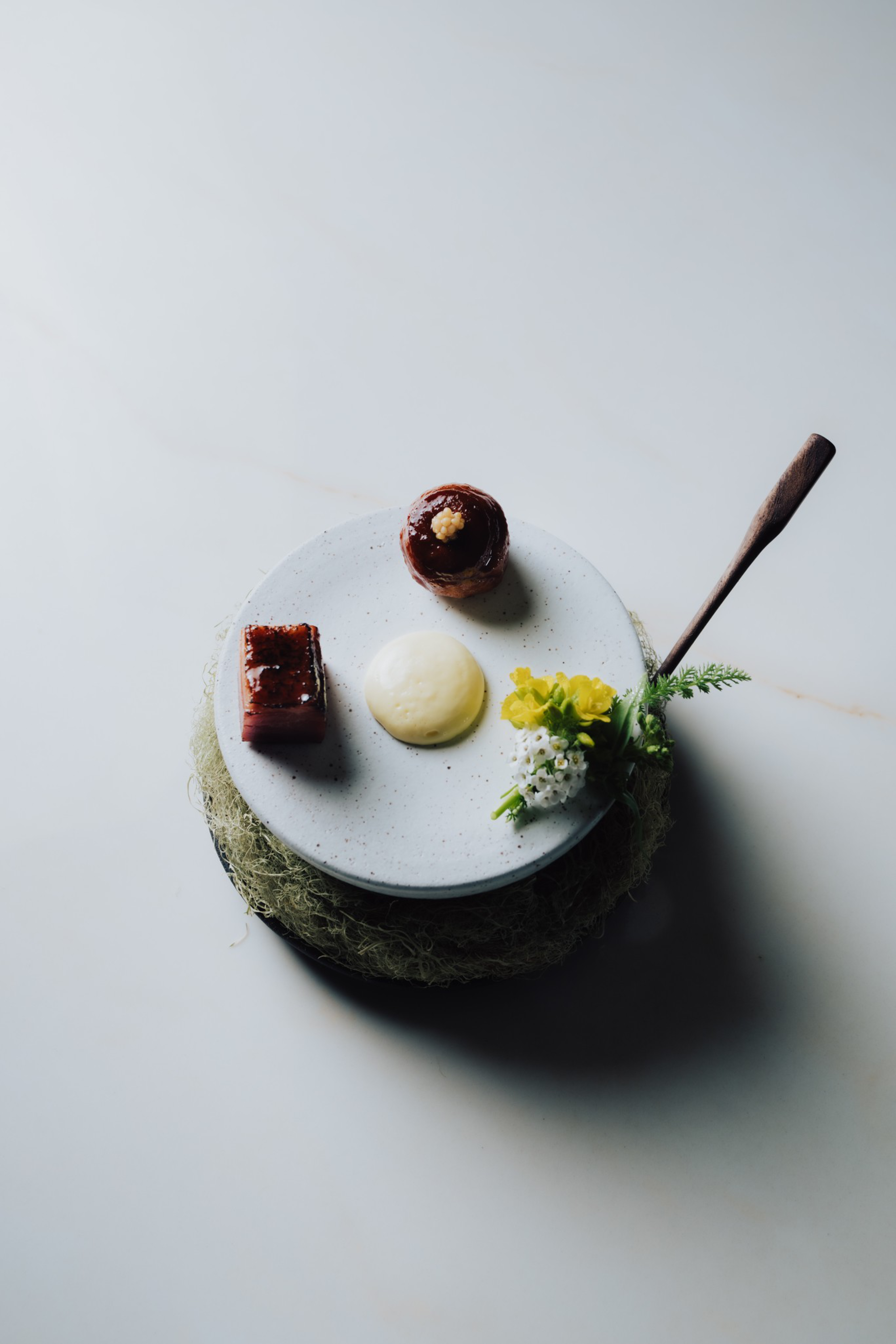 A white plate holds three small food items—a cube of glazed meat, a round meat portion with garnish, and a dollop of creamy sauce—alongside a small floral garnish.