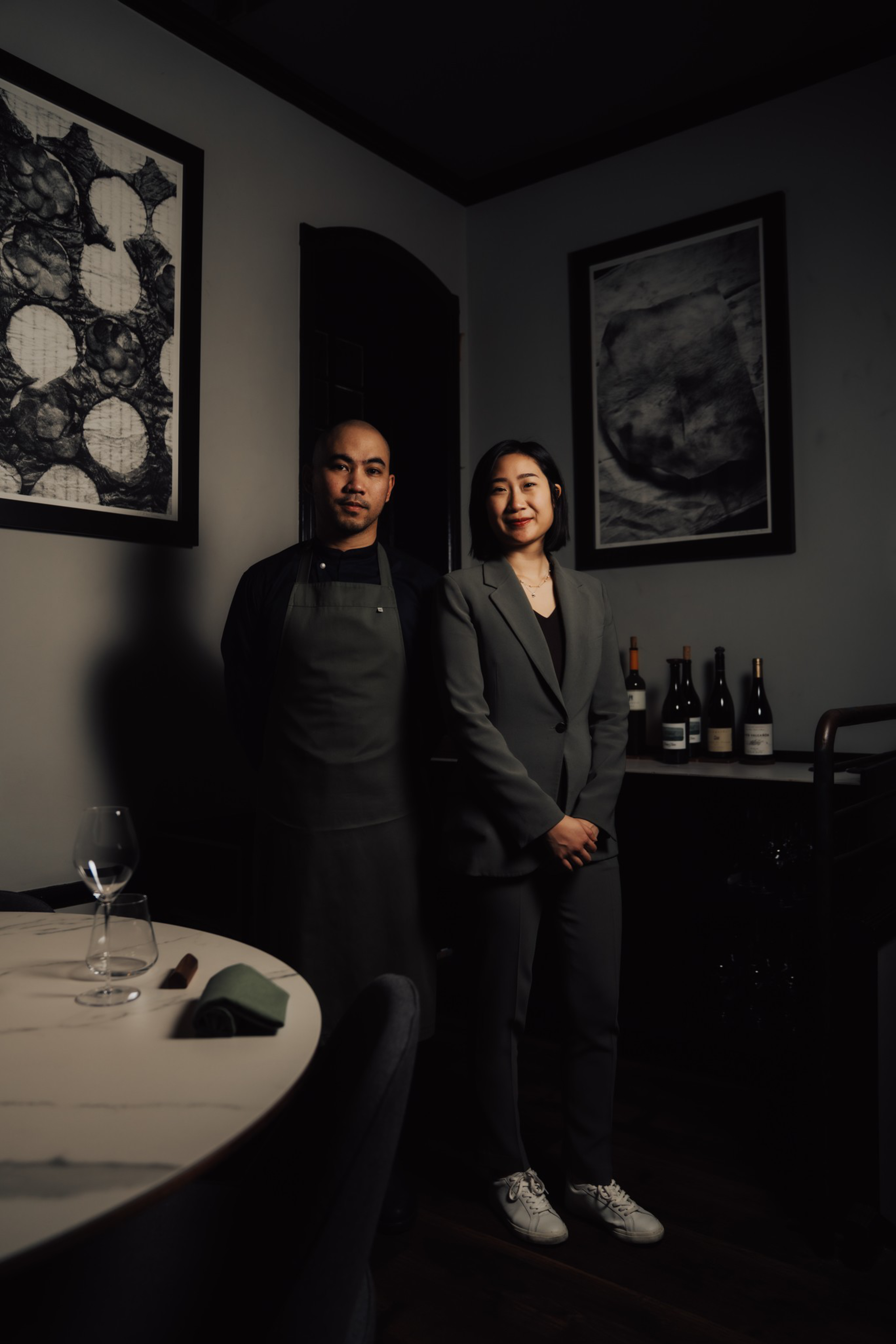 A man in a black apron and a woman in a gray suit stand side by side in a dimly lit room with abstract art and wine bottles in the background.
