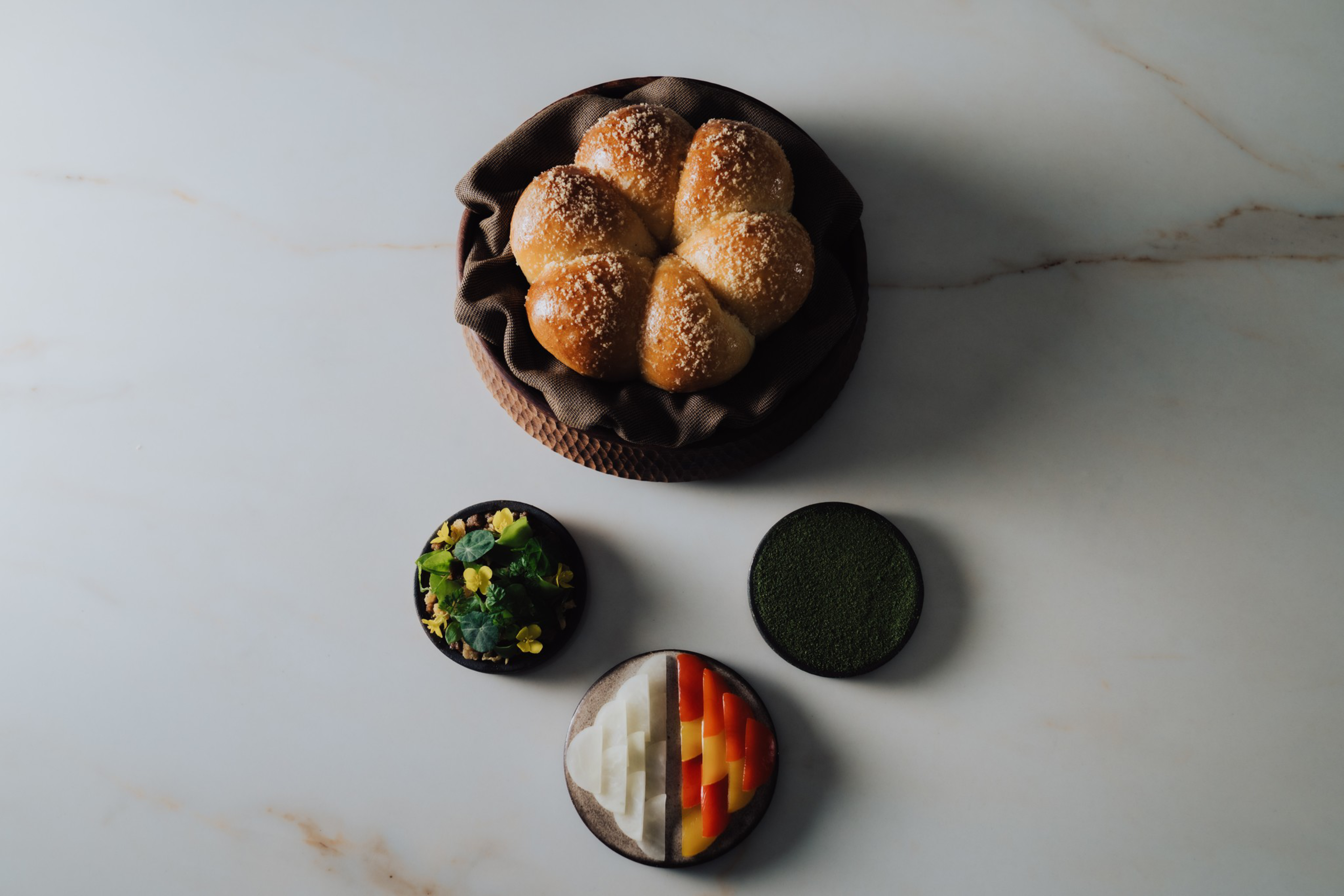 A basket with five round bread rolls topped with sugar is surrounded by three small dishes containing herbs, green powder, and sliced colorful vegetables.