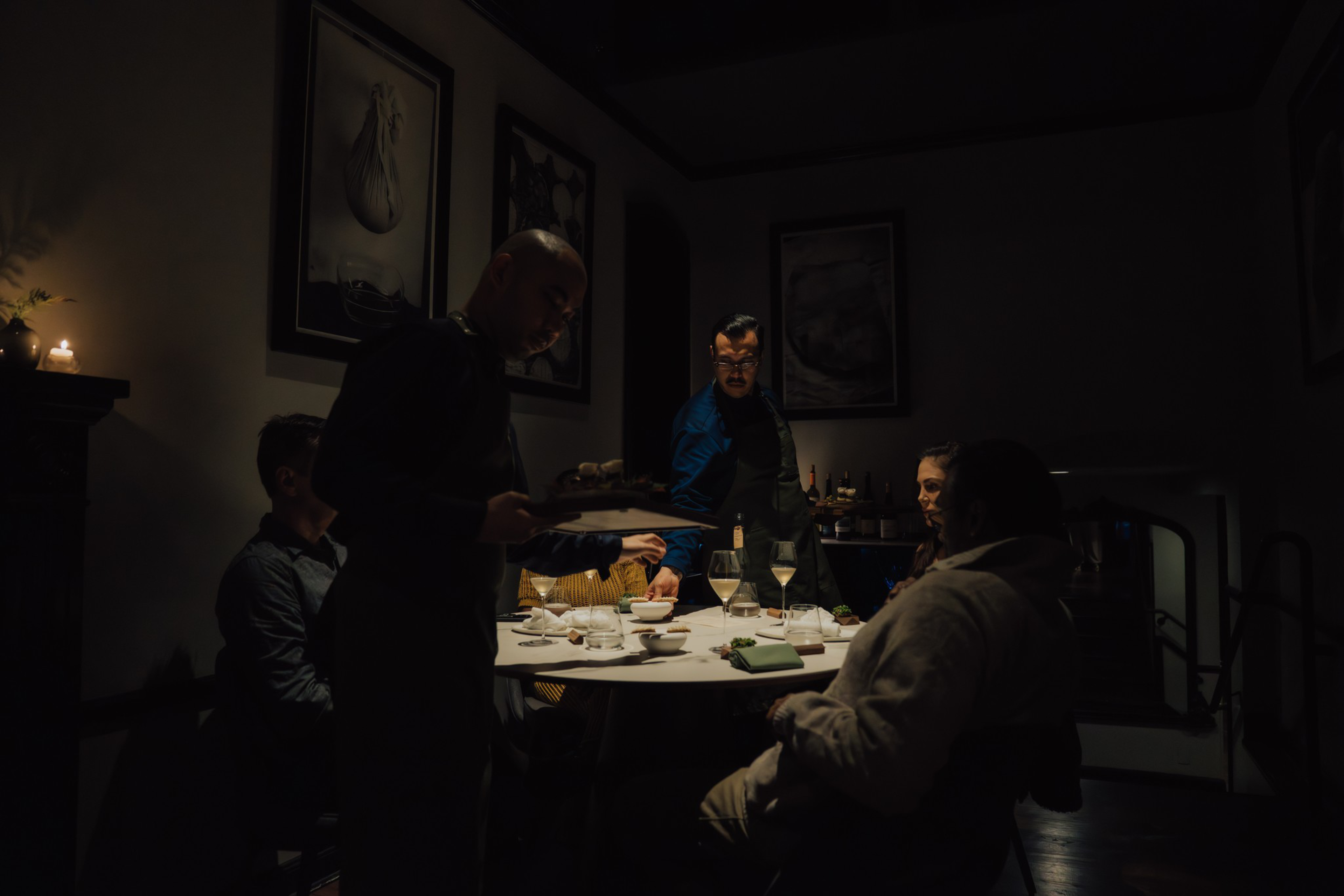 Four people sit around a dimly lit table with wine glasses and dishes as a server brings a plate, with dark walls and framed art in the background.