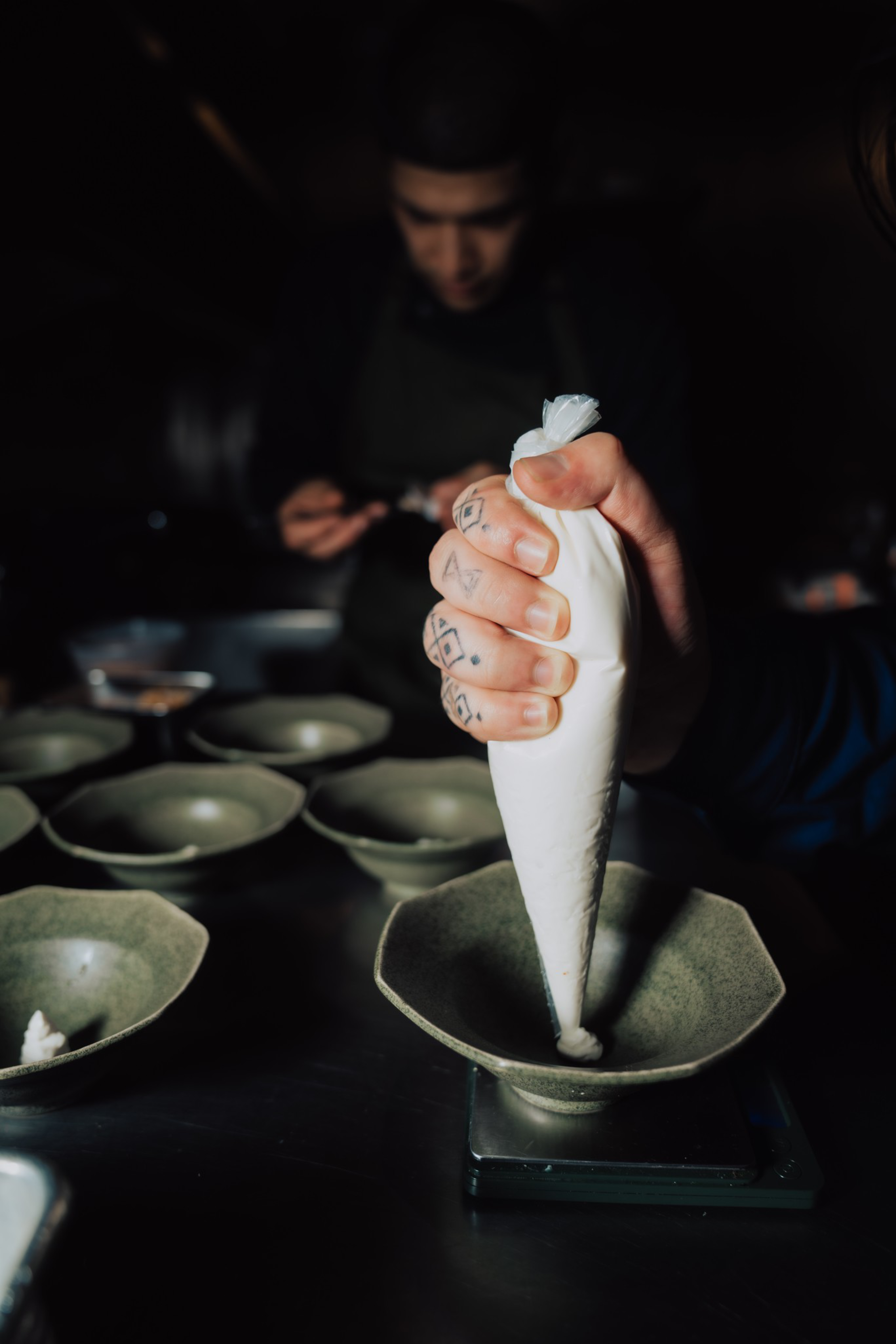 A tattooed hand is squeezing a white piping bag to fill a green bowl, with several similar bowls lined up on a dark surface in the background.