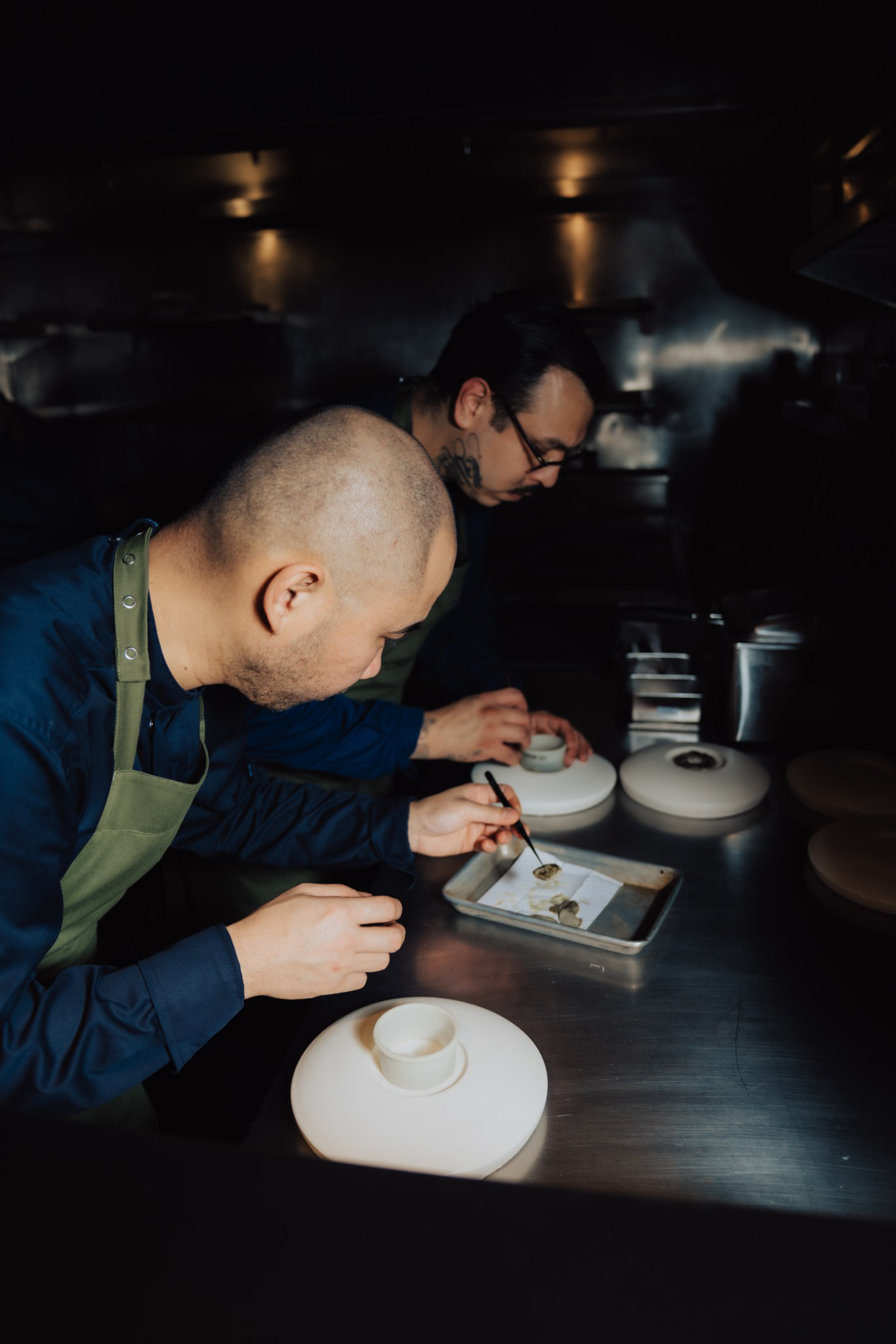 Two chefs wearing green aprons carefully plating food on white dishes in a dimly lit, professional kitchen.
