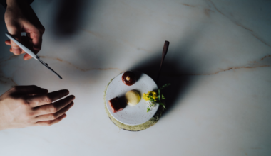 A small, elegant canapé topped with cream and peppercorns sits on a wooden board next to wild berries, leaves, and delicate white flowers.