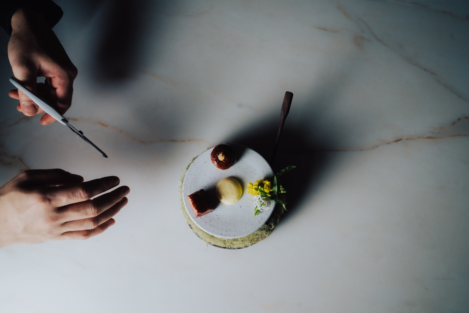 A small, elegant canapé topped with cream and peppercorns sits on a wooden board next to wild berries, leaves, and delicate white flowers.