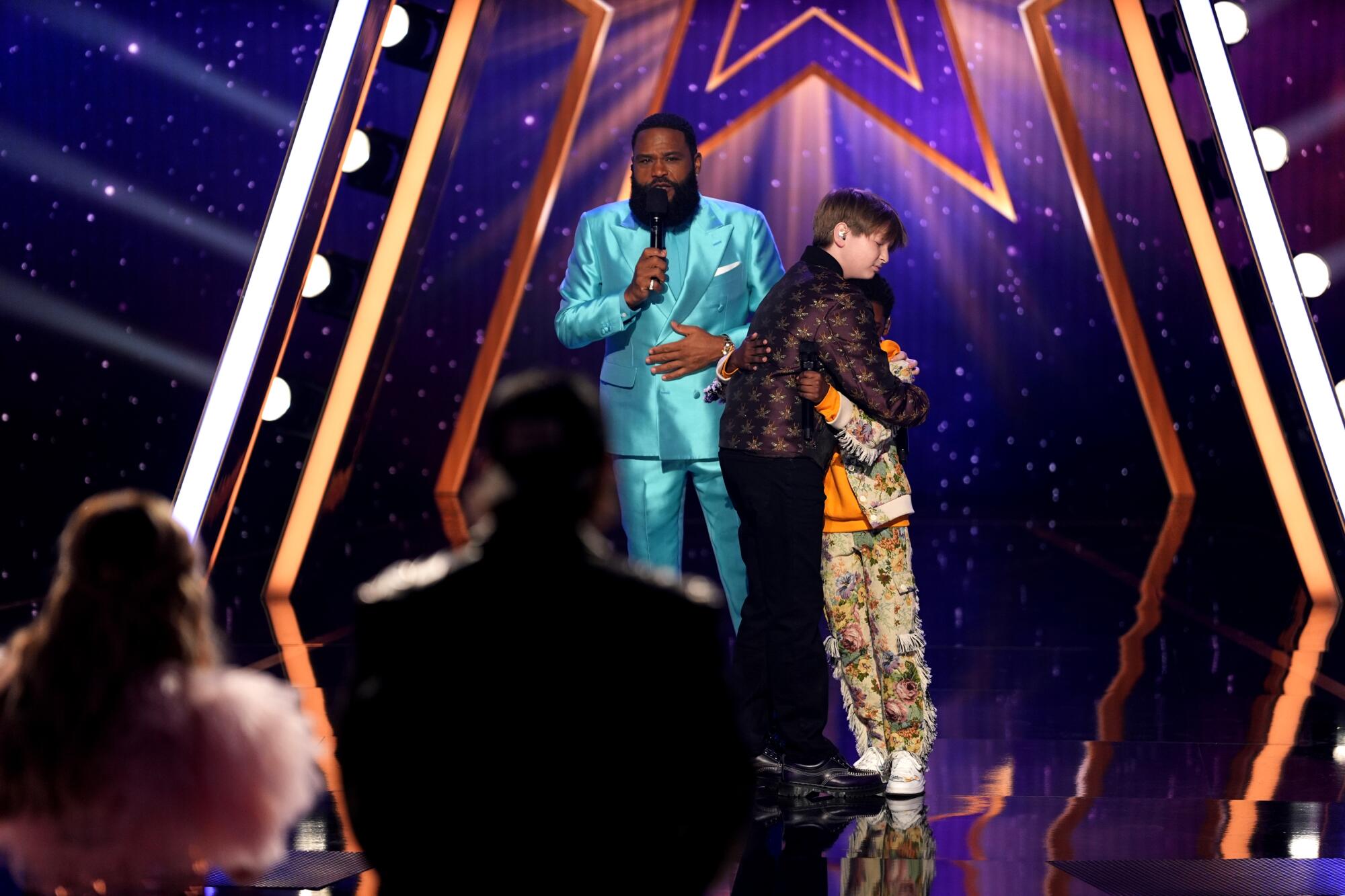 A man in a blue suit holding a microphone stands near two boys embracing on a stage.