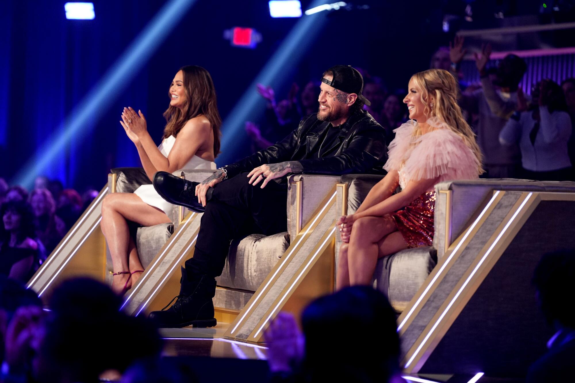 Three people sitting in chairs elevated on a stage with an audience surrounding them.