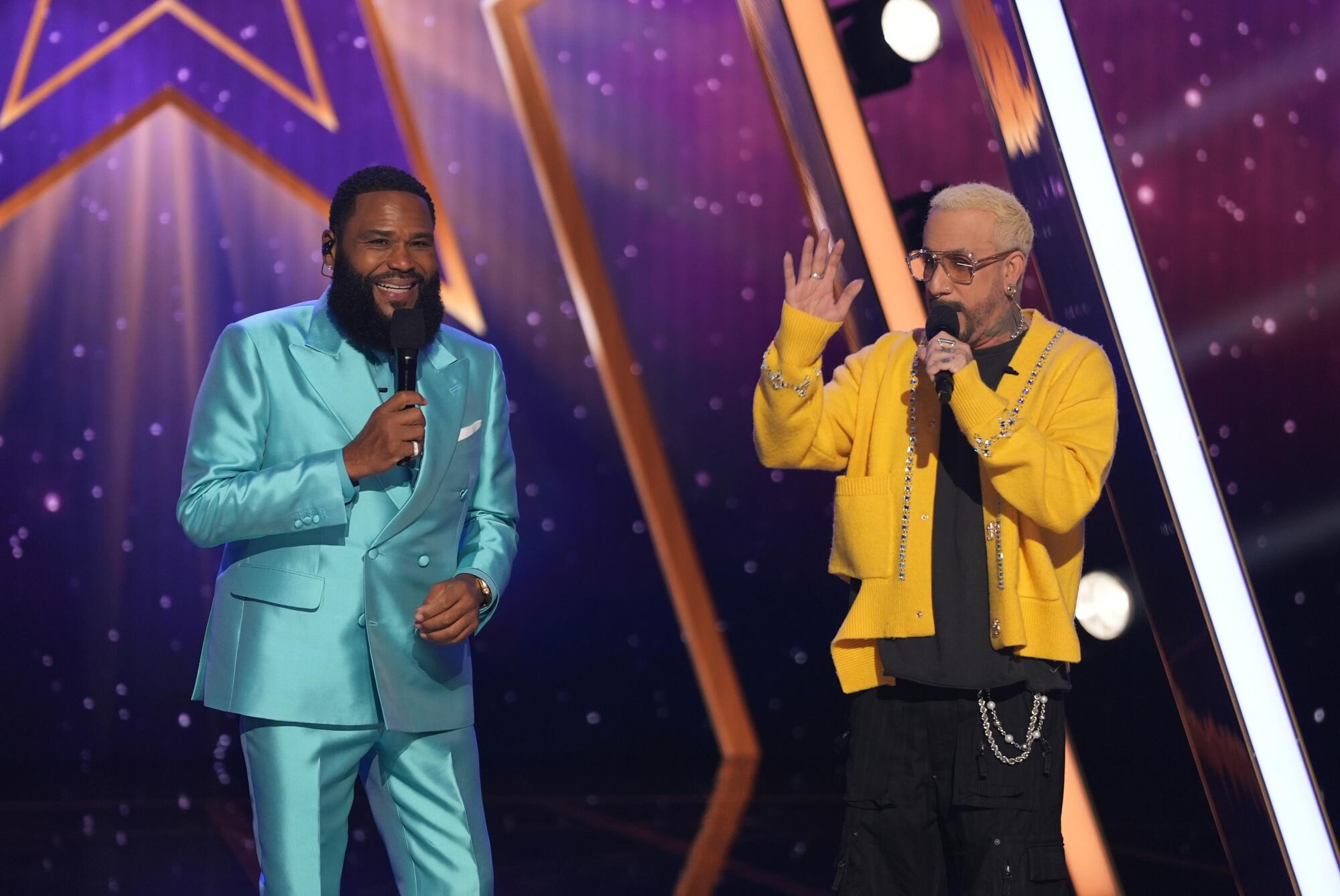 A man in a blue suit stands next to a man in a yellow jacket gesturing his hand.