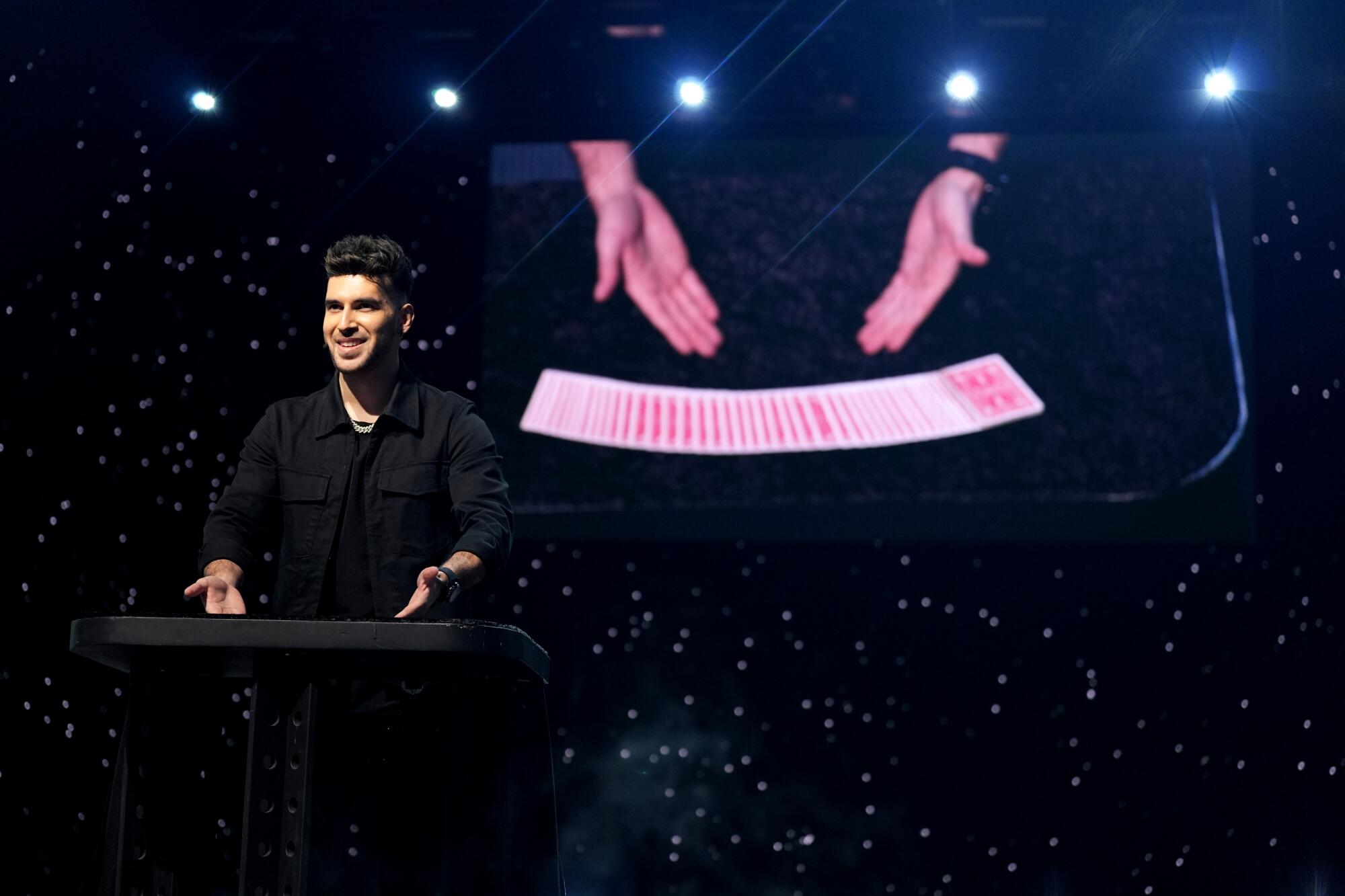 A man in a black shirt gestures at playing cards fanned out on a table. A screen behind him shows his hands and the cards.