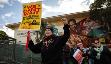 A woman holds a yellow sign reading "We Can't Wait" advocating school investment, surrounded by people and a mural outside Thurgood Marshall Academy.