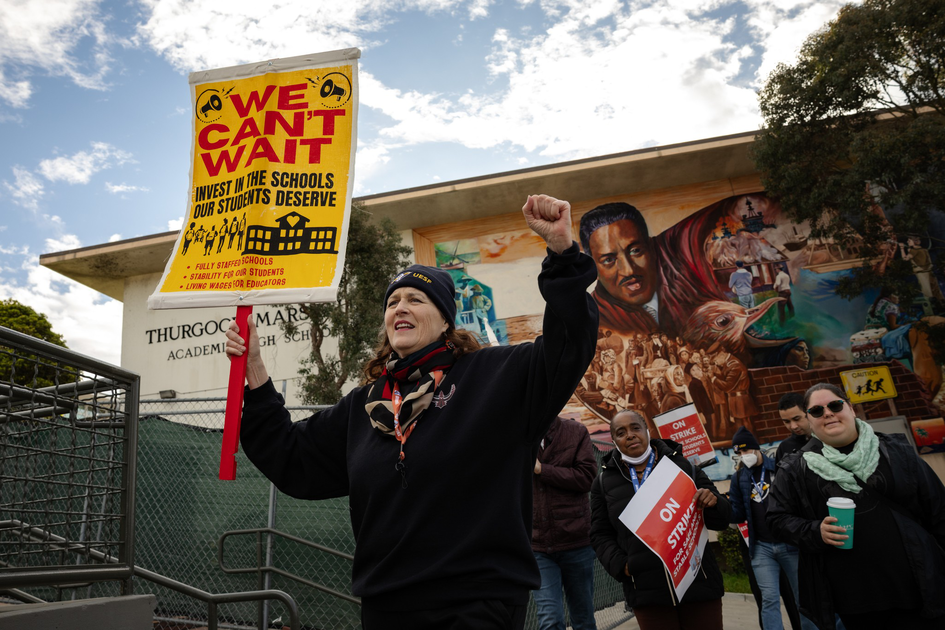 A woman holds a yellow sign reading "We Can't Wait" advocating school investment, surrounded by people and a mural outside Thurgood Marshall Academy.