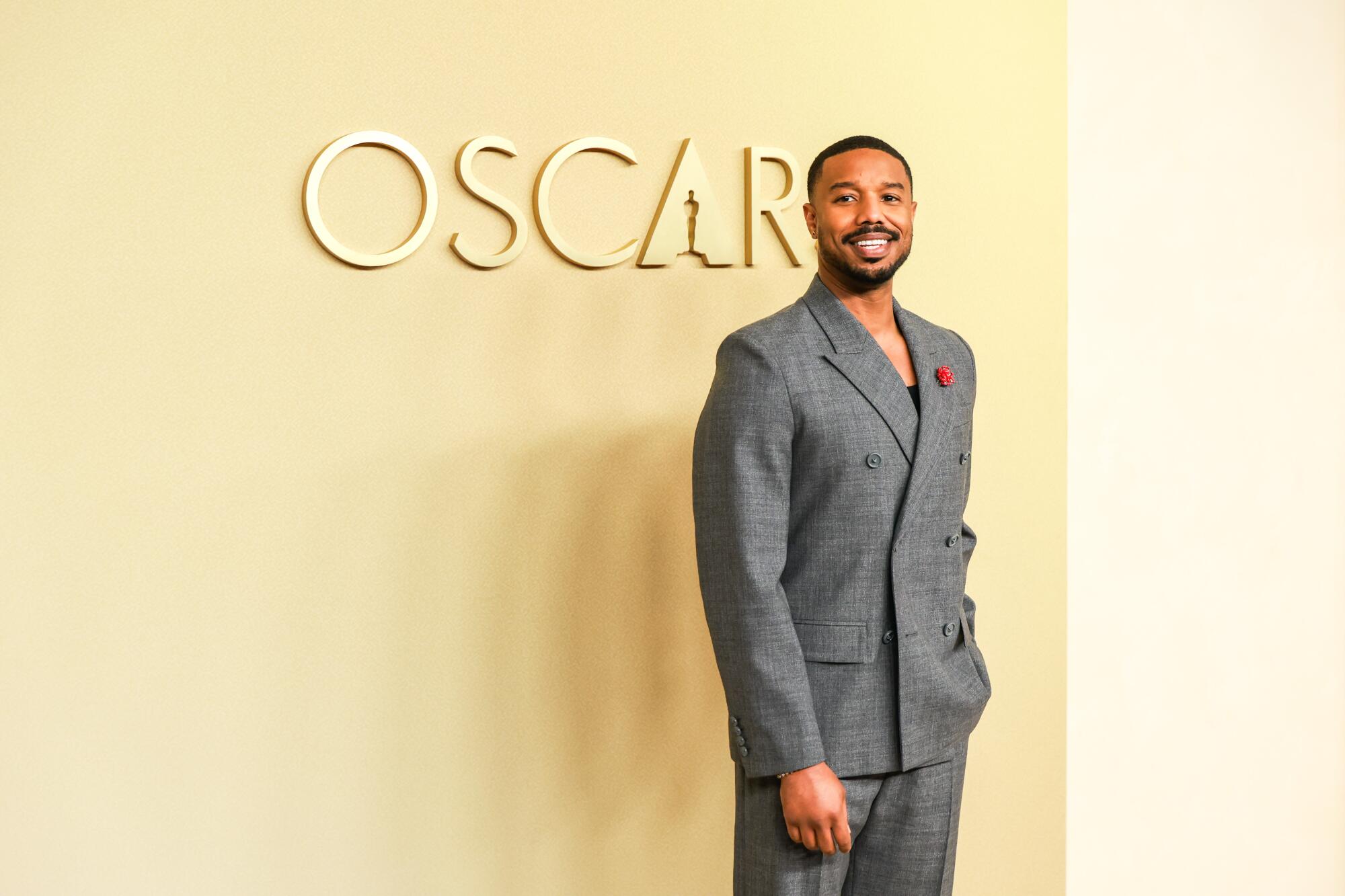 Michael B. Jordan, wearing a gray suit, poses in front of the Oscars logo. 
