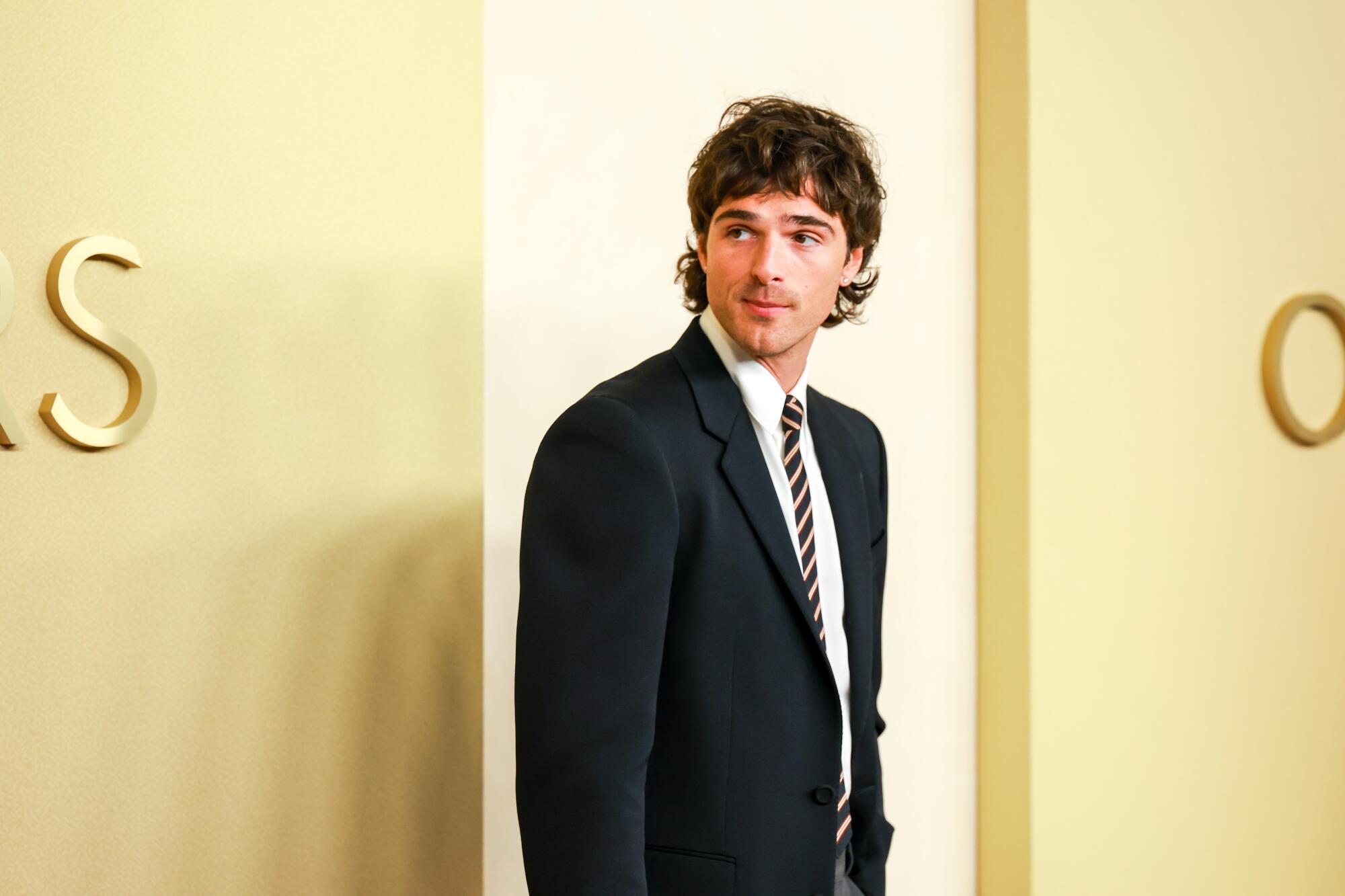 Jacob Elordi, wearing a suit and skinny tie, poses on the beige carpet. 