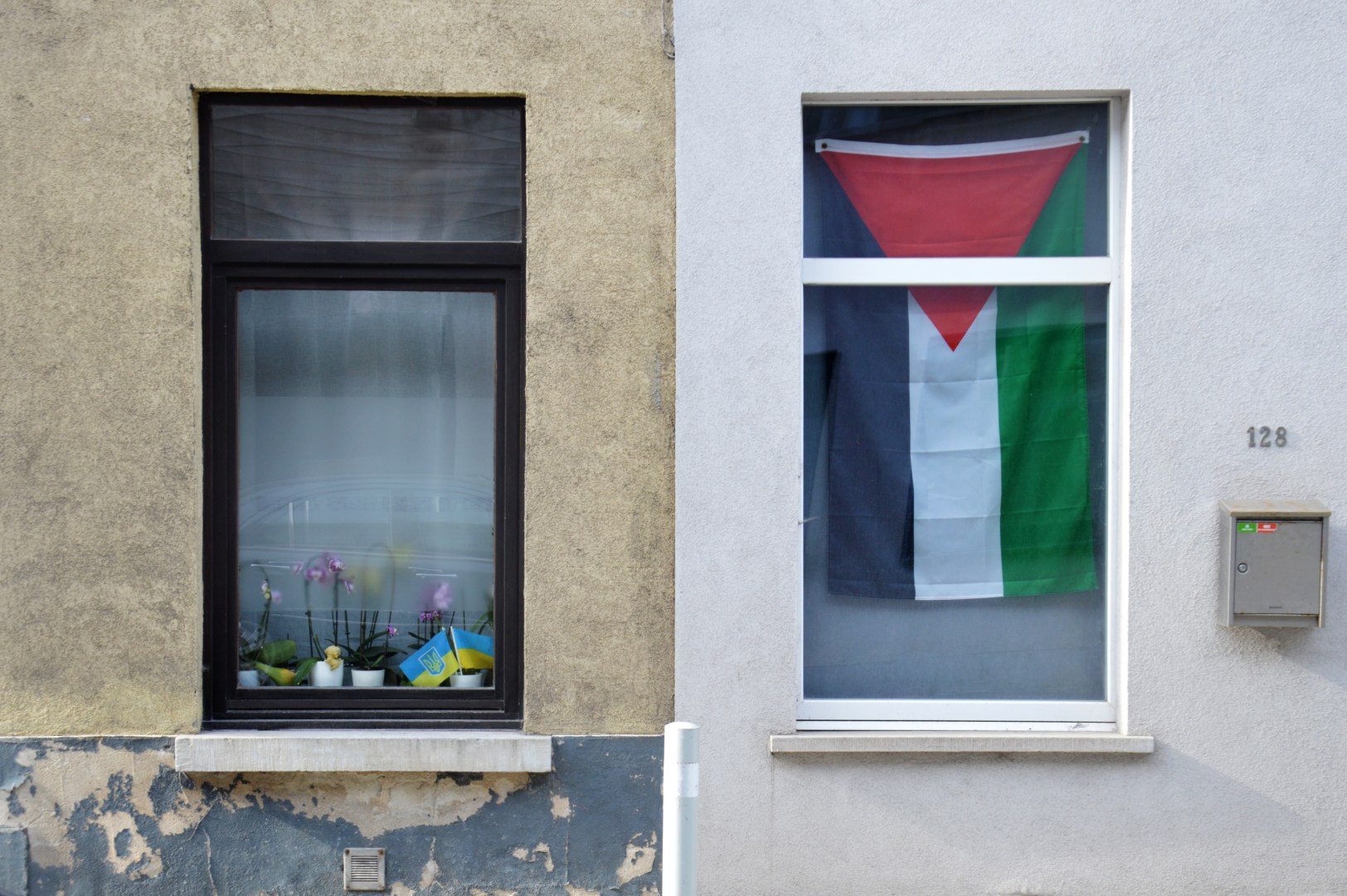 A photo of an apartment building with the Palestinian flag hanging inside a window on the right, with smaller Ukrainian flags displayed in a flower box on the left.