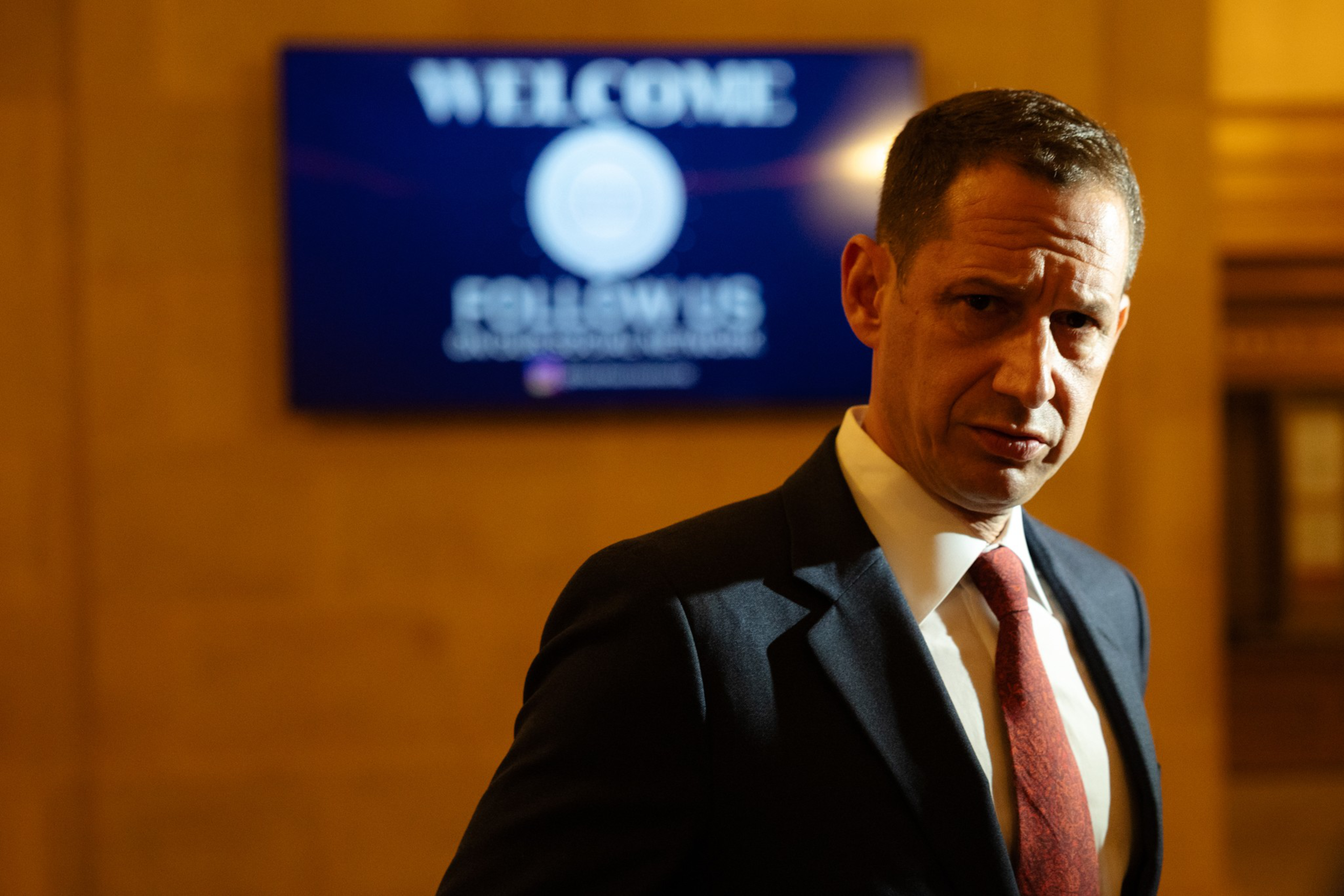 A man in a dark suit and red tie stands in front of a blurred screen displaying the words “WELCOME FOLLOW US.”