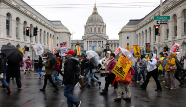 A group of people march on a wet street holding protest signs and umbrellas near a large domed building with "Franklin" street signs visible.