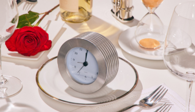 A silver clock on a white plate sits on a table with a red rose, wine bottle, forks, glasses, and an hourglass.