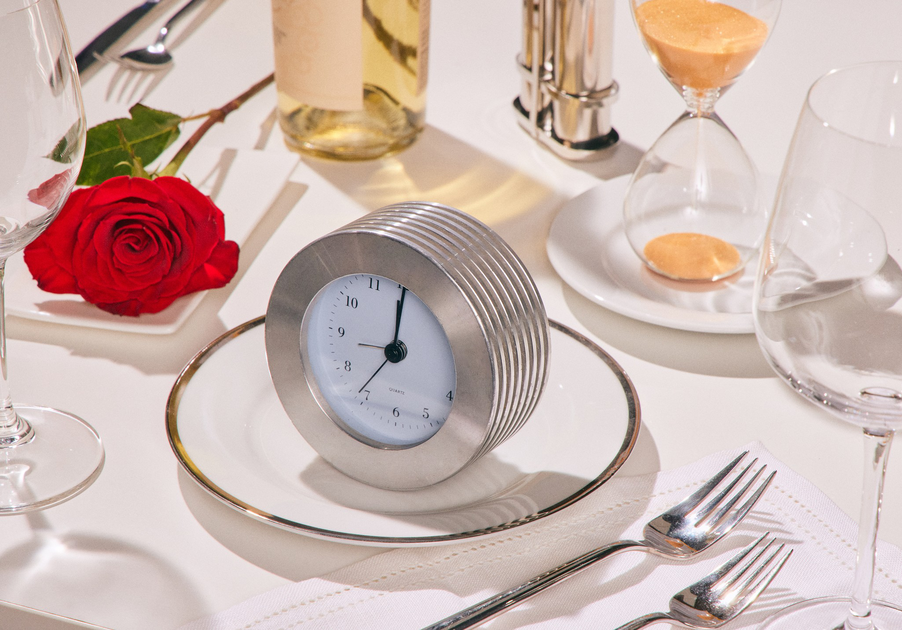 A silver clock on a white plate sits on a table with a red rose, wine bottle, forks, glasses, and an hourglass.