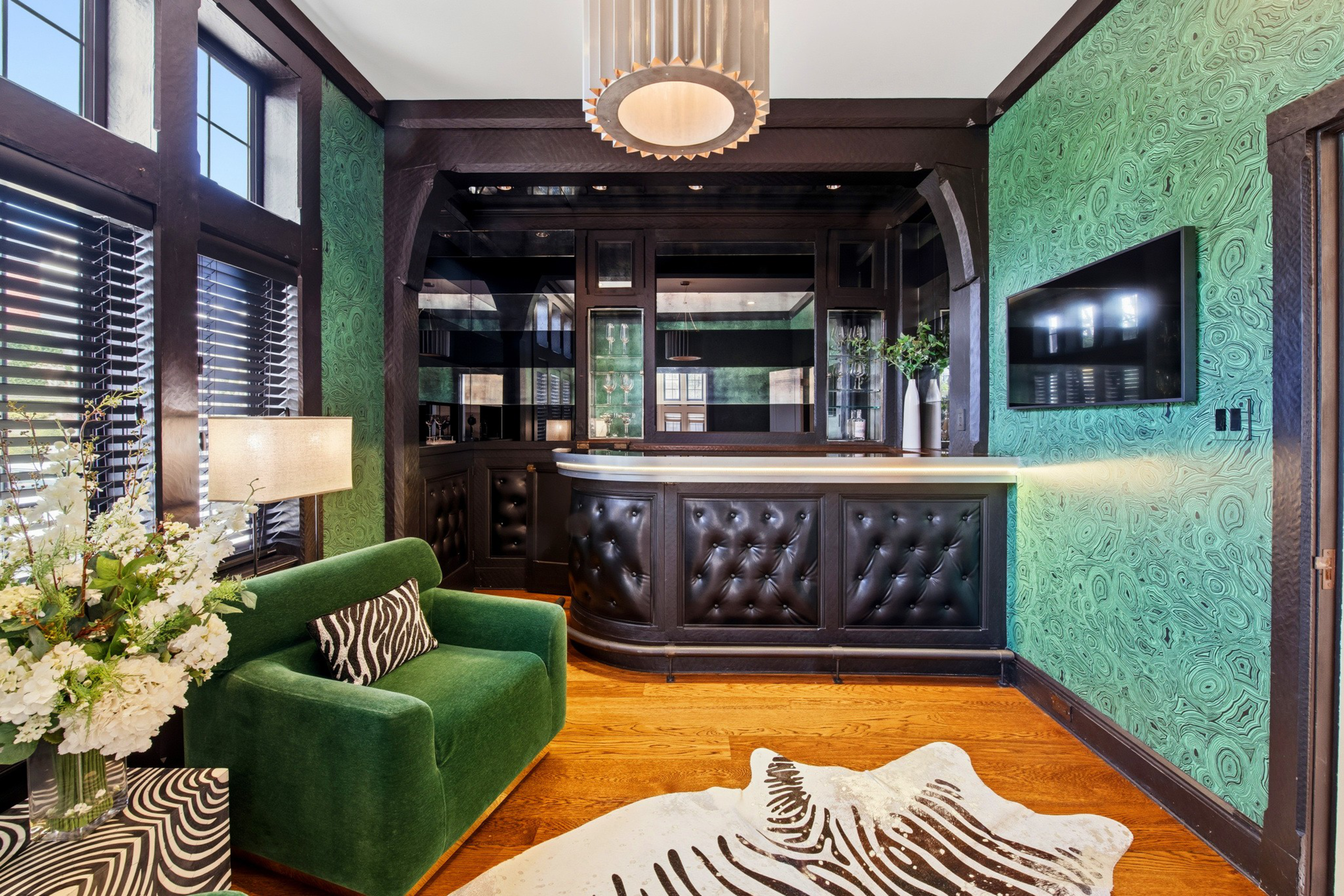 A small bar area features a dark tufted counter, green patterned walls, a green velvet chair, zebra print pillow and rug, and a wall-mounted TV.