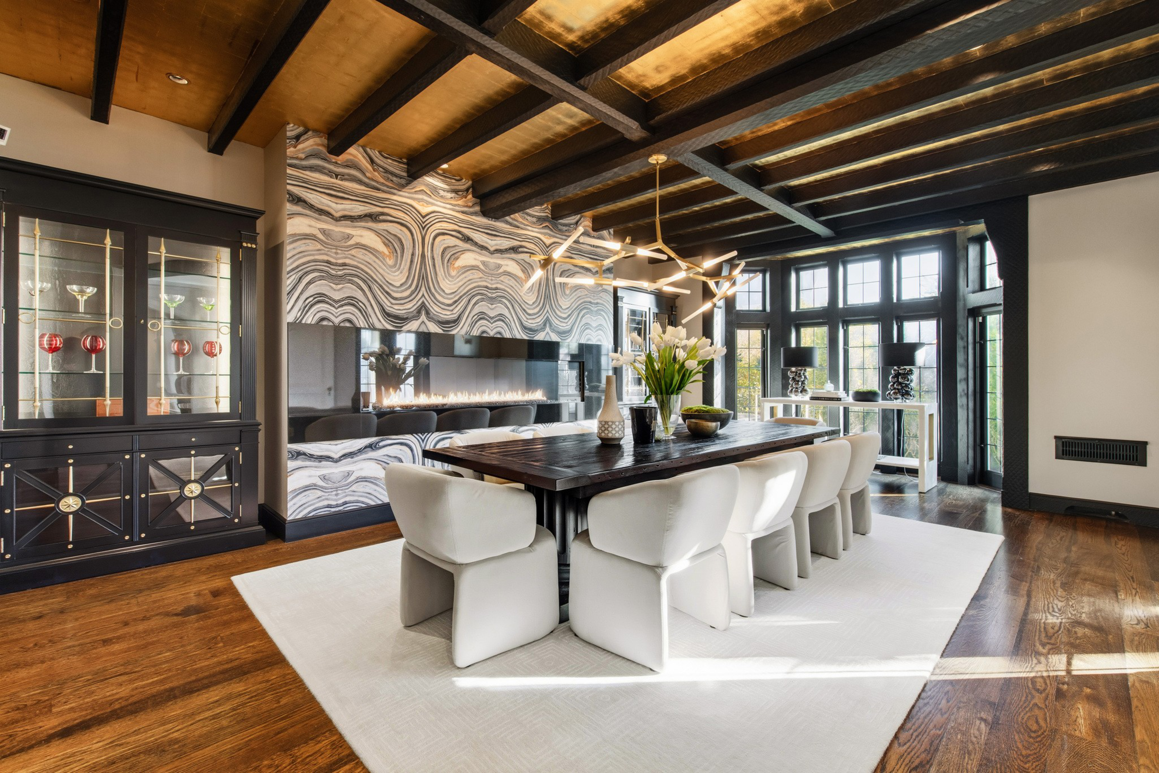 A modern dining room features a dark wood table surrounded by eight white upholstered chairs, a patterned stone accent wall with a built-in fireplace, and large windows.