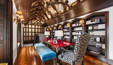 A long room with wooden vaulted ceiling, red glossy table, patterned chairs, blue bench, wall sconces, chandeliers, and bookshelves filled with books and decor.