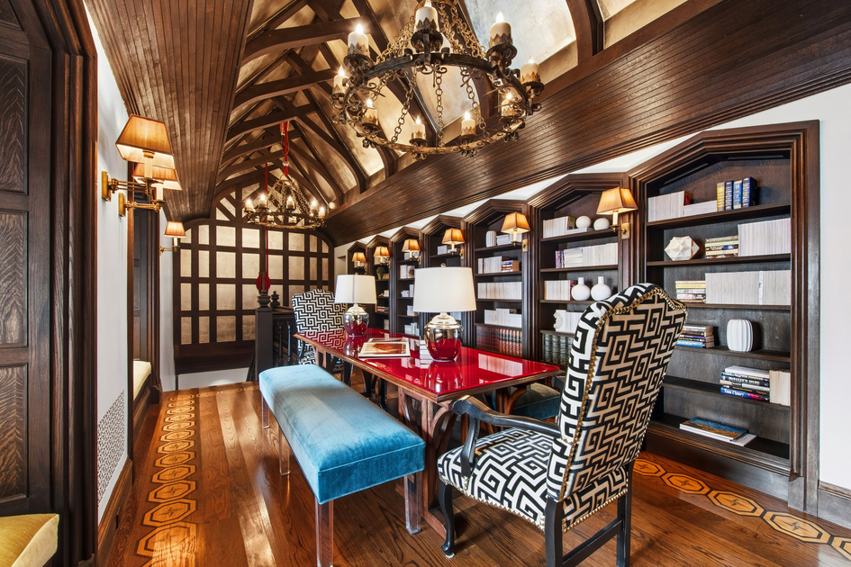 A long room with wooden vaulted ceiling, red glossy table, patterned chairs, blue bench, wall sconces, chandeliers, and bookshelves filled with books and decor.