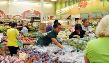 Several people shop for fresh produce in a grocery store, with fruits like pineapples, melons, and grapes visible under bright store lighting.