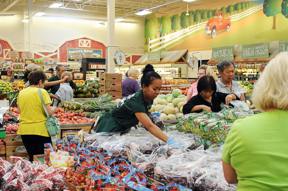 Several people shop for fresh produce in a grocery store, with fruits like pineapples, melons, and grapes visible under bright store lighting.