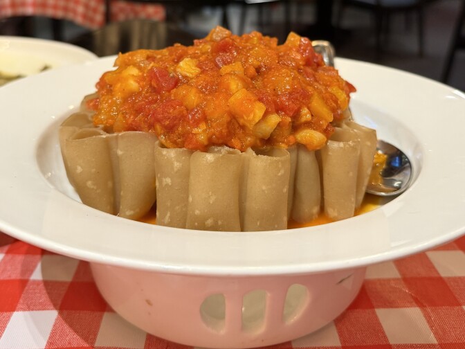 A white plate contains a circle of thick cylindrical noodles, on top of which is a red stew