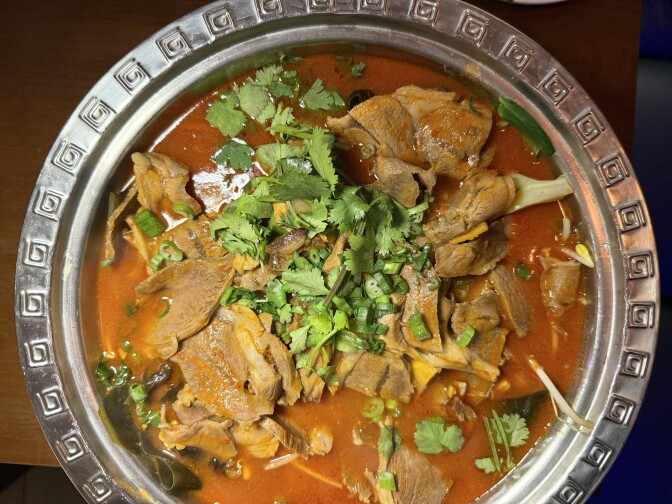 A metal bowl with decorative edges contains a tomato-based liquid filled with brown pieces of meat and a green vegetable garnish