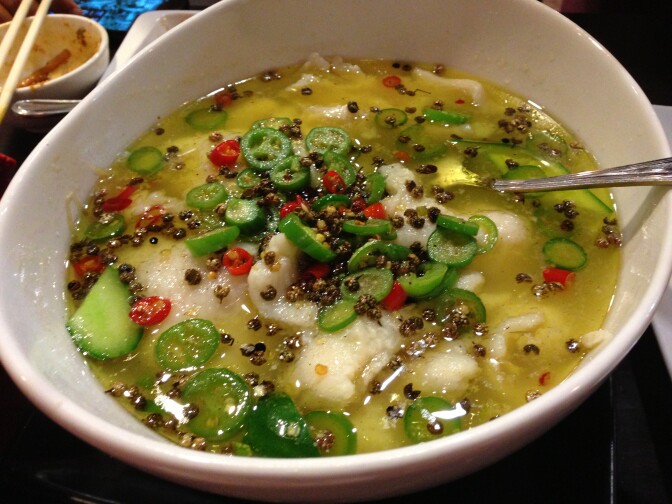 A white oval bowl contains a yellow broth which holds white pieces of meat, green and red vegetables and peppercorns