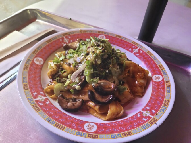 A white plate with a traditional red and gold decoration around the side holds a dish of sauced noodles covered with stir-fried vegetables