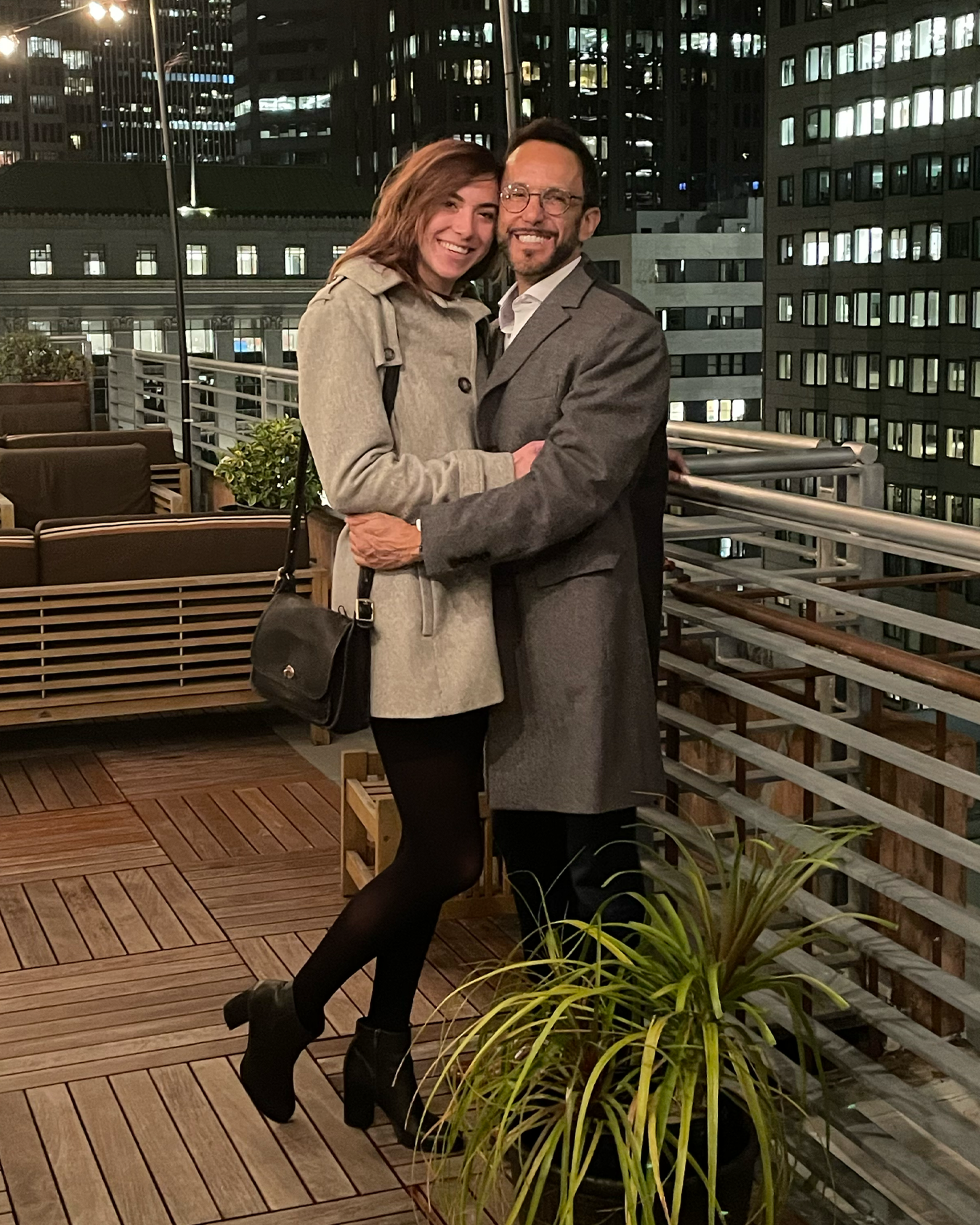 A man and woman dressed in coats embrace and smile on a rooftop patio at night with city buildings lit in the background.