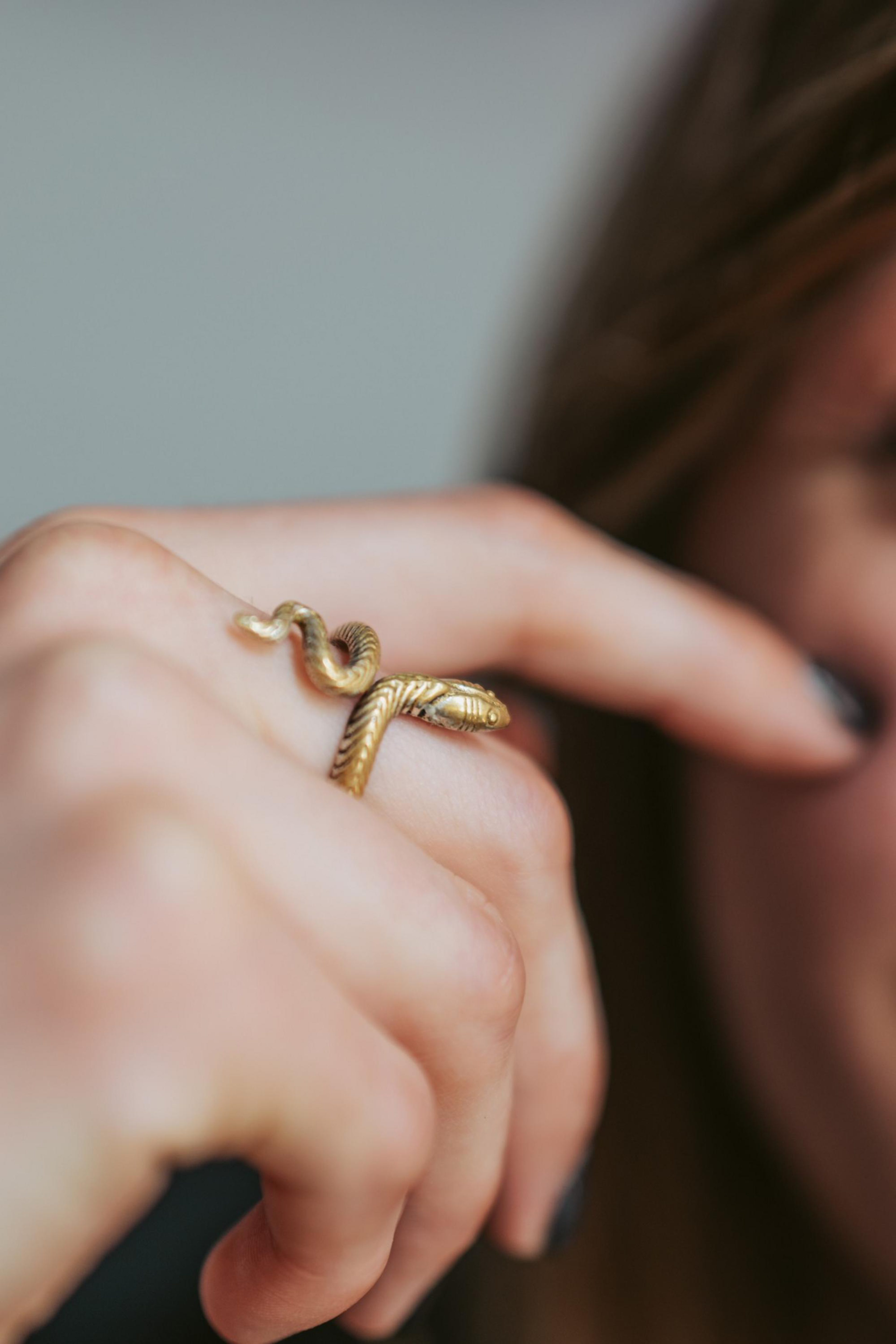 A close-up of a hand wearing a gold snake-shaped ring coiled around the finger, with part of a face blurred in the background.