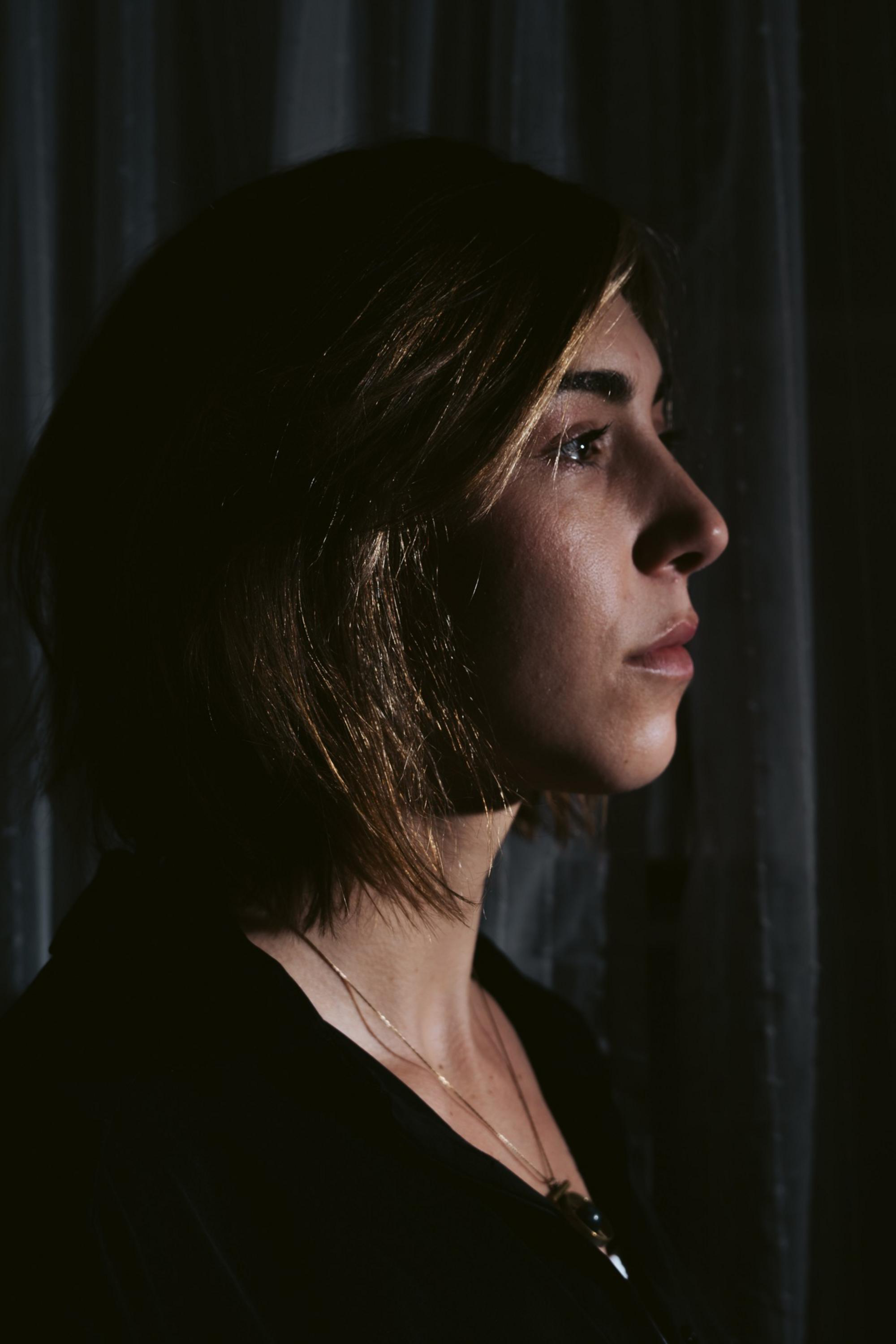 A woman with shoulder-length hair faces right in dim light, her profile softly illuminated against a dark, curtain backdrop.