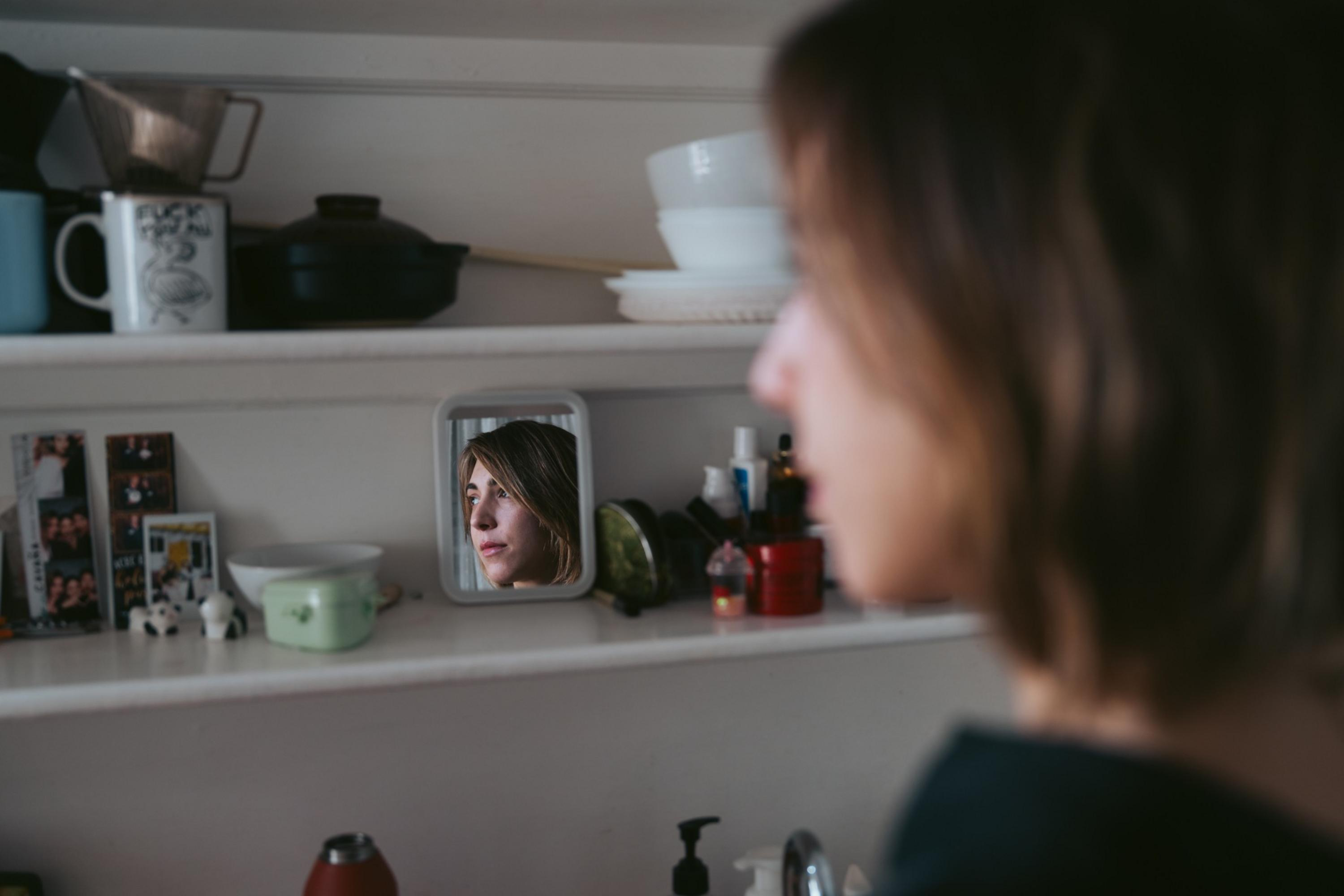 A woman’s face is reflected in a small mirror on a shelf filled with cups, bowls, photos, and various bottles.