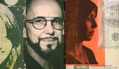 A man and woman dressed in coats embrace and smile on a rooftop patio at night with city buildings lit in the background.