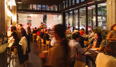 A busy restaurant with warm lighting shows diners seated at tables and a bar, with large windows revealing a nighttime street view outside.