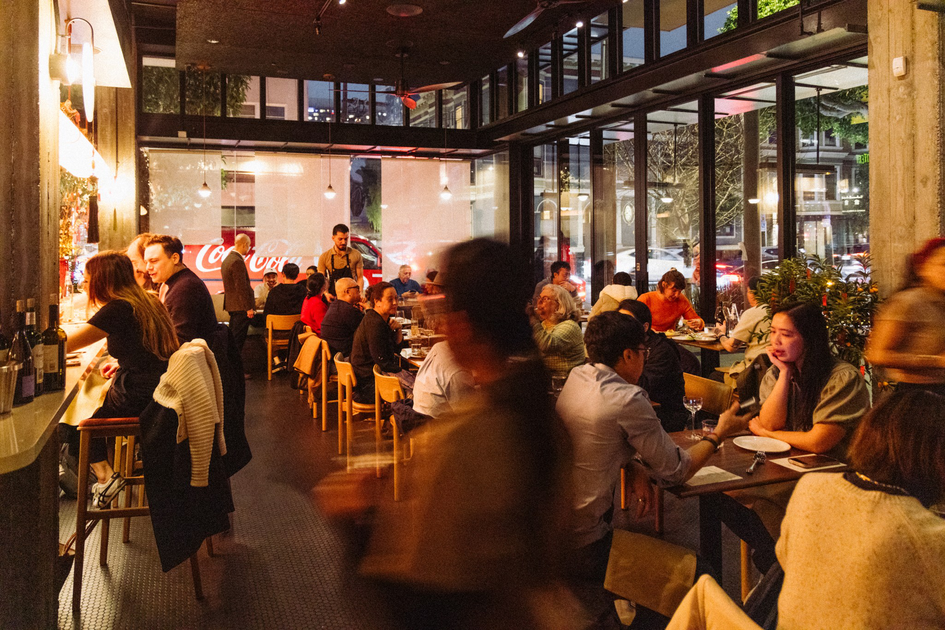 A busy restaurant with warm lighting shows diners seated at tables and a bar, with large windows revealing a nighttime street view outside.