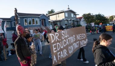A group of people gather outdoors near houses, with one holding a large sign that reads, "DR. SU, EDUCATORS NEED A FAIR CONTRACT NOW!"