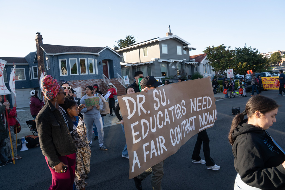 A group of people gather outdoors near houses, with one holding a large sign that reads, "DR. SU, EDUCATORS NEED A FAIR CONTRACT NOW!"